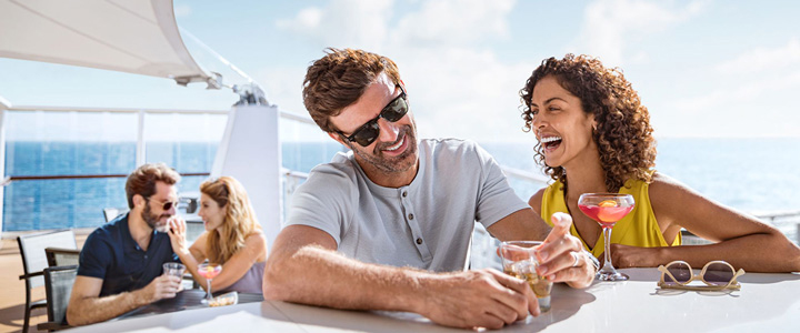 Lachendes Pärchen sitzt bei einem Glas Alkohol an einem Tisch an Bord eines Kreuzfahrtschiffes bei schönem Wetter; weiteres Pärchen im Hintergrund.